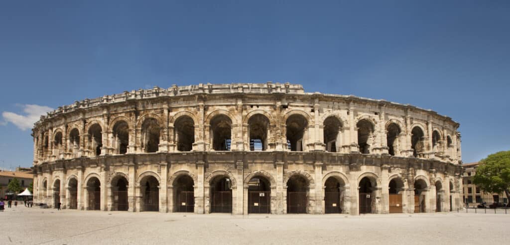 Arènes de Nîmes, amphithéâtre romain antique en pierre beige sous un ciel bleu clair, avec des portes d'entrée fermées au rez-de-chaussée.