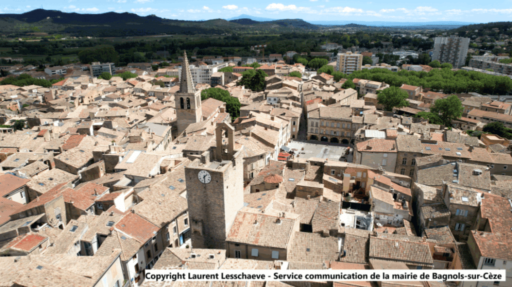 Vue aérienne de Bagnols-sur-Cèze. Centre historique, toits de tuiles, beffroi à horloge et clocher d'église sous ciel bleu.