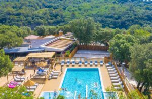 Vue aérienne d'un camping avec piscine extérieure entourée de transats, terrasse en bois et d'un terrain de pétanque, au pied d'une colline boisée.