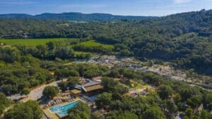 Vue aérienne d'un camping provençal : piscine, terrasses, et bâtiments, au milieu d'une forêt dense. Une rivière rocheuse, des vignobles et des collines boisées sont visibles en arrière-plan.
