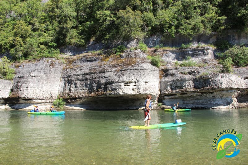 Paddleboarder et kayaks sur la rivière Cèze devant des falaises calcaires boisées. Logo "CÈZE CANOËS".