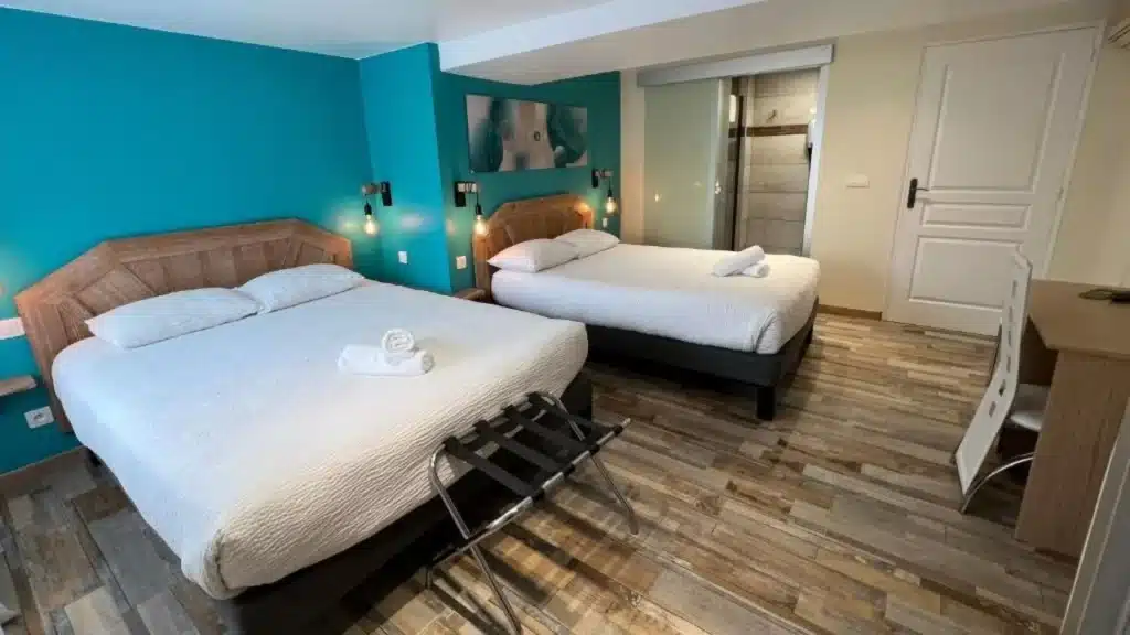 Chambre d'hôtel lumineuse avec deux lits, mur turquoise vif, têtes de lit en bois texturé, serviettes roulées et porte coulissante en verre menant à la salle de bain.