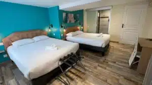 Chambre d'hôtel lumineuse avec deux lits, mur turquoise vif, têtes de lit en bois texturé, serviettes roulées et porte coulissante en verre menant à la salle de bain.