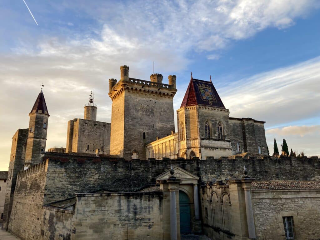 Château médiéval en pierre au coucher du soleil, avec une tour crénelée, un toit de tuiles colorées et un avion traçant une condensation dans le ciel bleu.