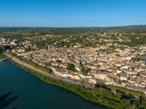 Cité historique provençale vue du ciel, avec ses toits tuilés et son clocher, bordant un grand fleuve bleu. Routes et forêts environnantes.