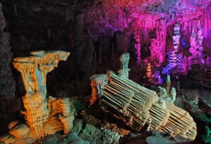 Intérieur d'une grotte avec des formations de stalactites et stalagmites illuminées en rose, violet et orange vif, sentier visible.