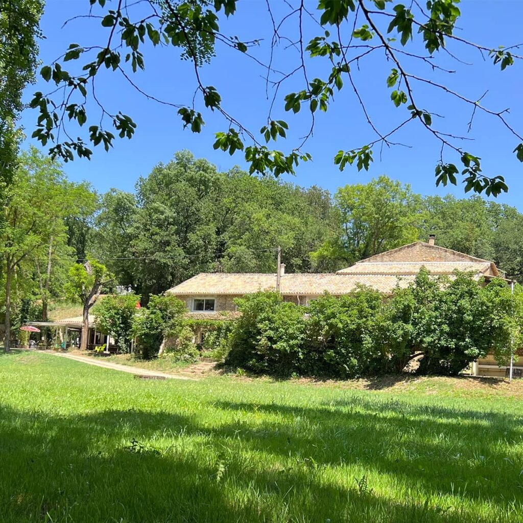 Maison en pierre traditionnelle avec toit de tuiles sous un ciel bleu éclatant, entourée d'une végétation luxuriante et d'un champ d'herbe verte.