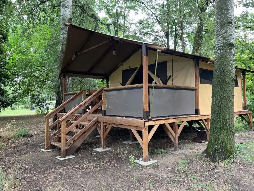 Tente safari glamping sur terrasse bois surélevée. Cabane en toile beige, escaliers en bois rustiques, au milieu d'une forêt luxuriante.