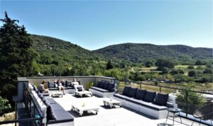 Terrasse moderne avec mobilier en palettes et coussins gris, des personnes se détendent avec vue sur des collines verdoyantes et ensoleillées.