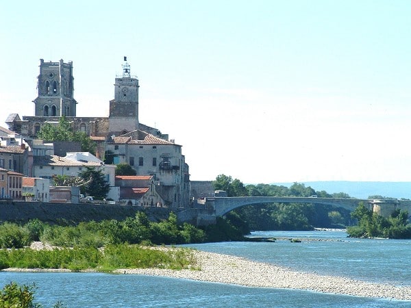 Vue d'un village provençal surélevé avec une cathédrale aux deux tours dominant les toits, surplombant une rivière avec un pont en pierre à arche unique.
