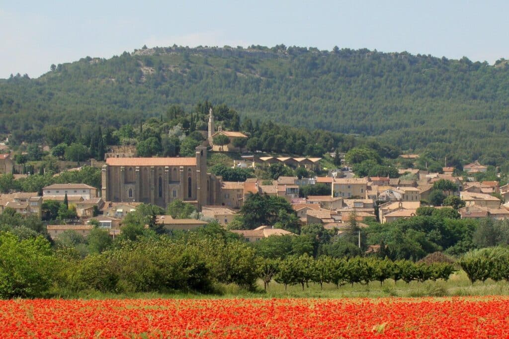 Champ de coquelicots rouges au premier plan, village provençal aux toits de tuiles et église en pierre, dominé par une colline boisée.