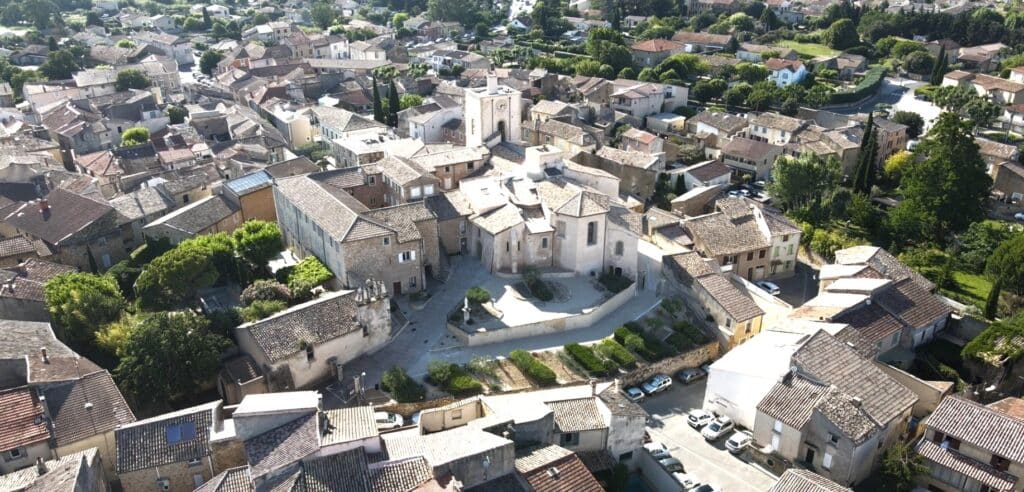 Vue aérienne d'un village provençal typique avec maisons en tuiles, une église centrale avec clocher-tour et une place pavée ensoleillée.