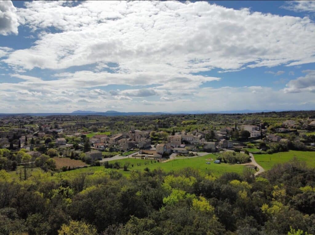 Vue aérienne d'un petit village provençal avec des toits de tuiles, entouré de végétation printanière luxuriante sous un ciel bleu parsemé de gros nuages blancs.