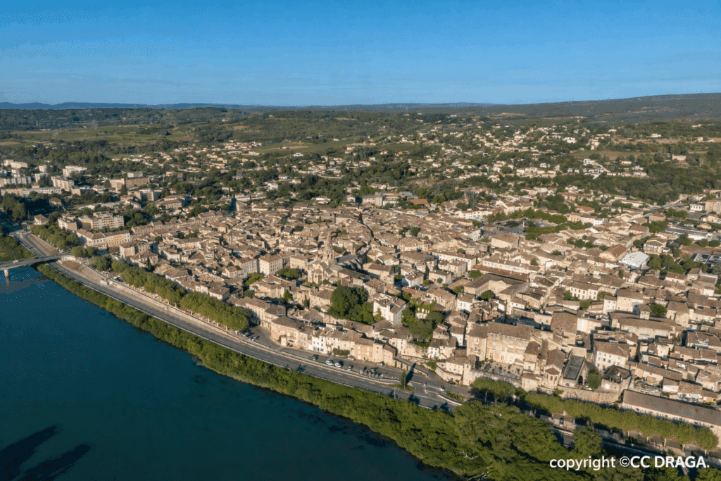 Vue aérienne d'une vieille ville provençale le long d'un large fleuve. Maisons aux toits de tuiles, cernées par les collines et la verdure.