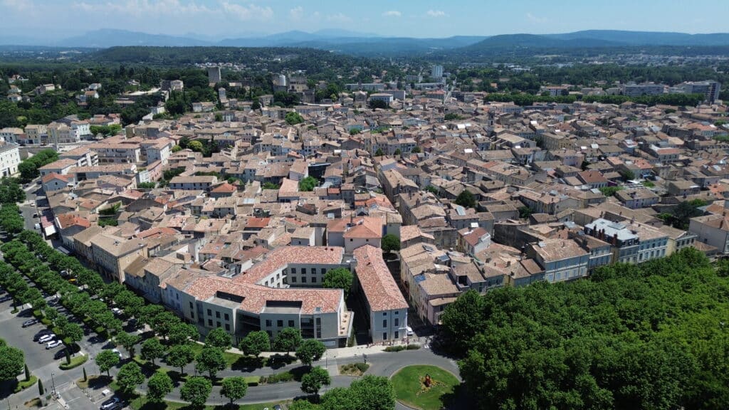 Vue aérienne d'une ville provençale dense aux toits de tuiles rouges, entourée de verdure et de collines bleutées en arrière-plan.