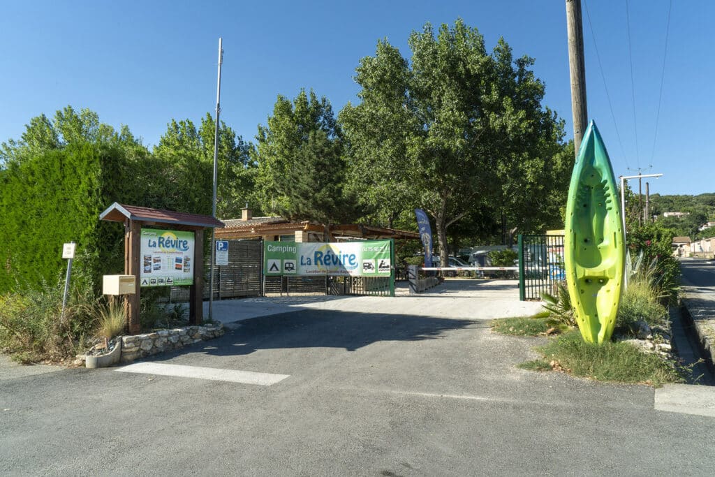 Entrée du Camping La Révire, location de canoës-kayaks. Panneaux, barrière et grand kayak décoratif vert vif sous le soleil.