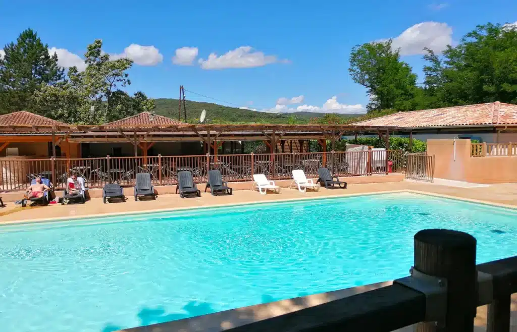 Piscine turquoise ensoleillée avec terrasse ombragée et transats. Deux personnes se relaxent face aux collines verdoyantes sous un ciel bleu d'été.