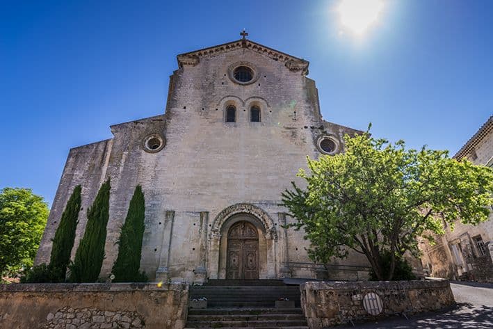 Ancienne église en pierre de Provence baignée de soleil. Escalier montant à la porte romane, encadrée par des cyprès et un grand arbre feuillu.