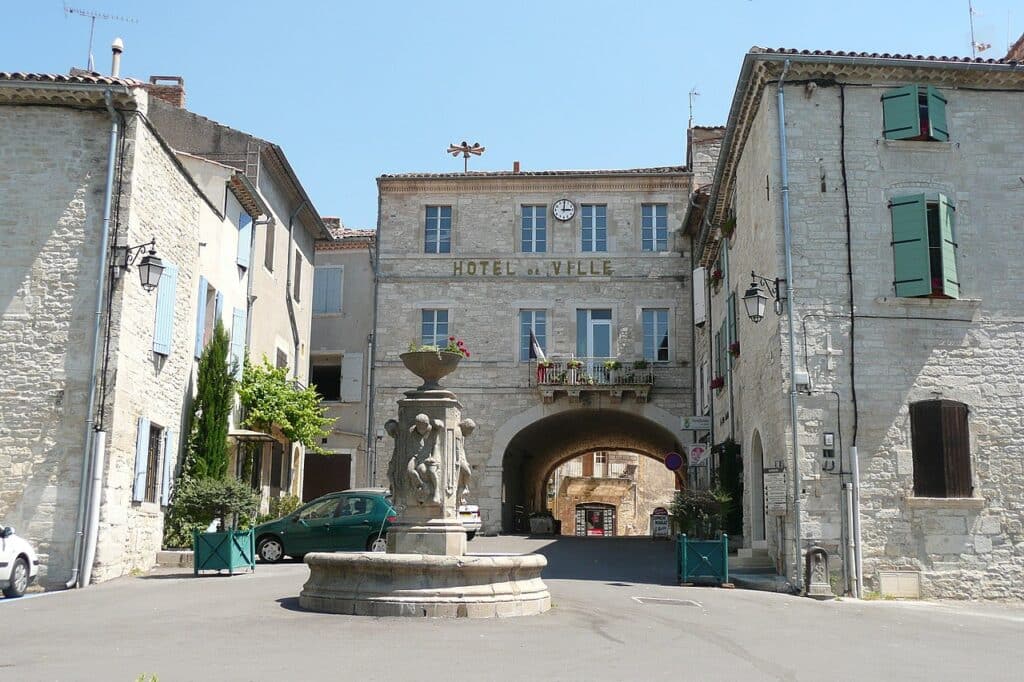 Place ensoleillée d'un village provençal. Fontaine centrale devant l'Hôtel de Ville en pierre avec son arche et son horloge.
