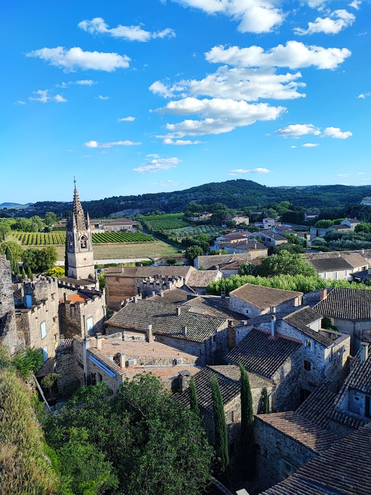 Paysage de Provence: toits de tuiles d'un village médiéval, clocher pointu, remparts et vignobles verdoyants sous un ciel bleu éclatant.