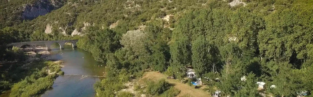 Camping ombragé en bord de rivière avec un pont de pierre historique dans une vallée verdoyante ensoleillée.
