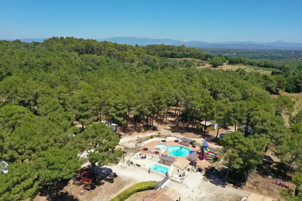 Vue aérienne d'un camping avec piscine et toboggans niché dans une vaste forêt sous un ciel bleu radieux.