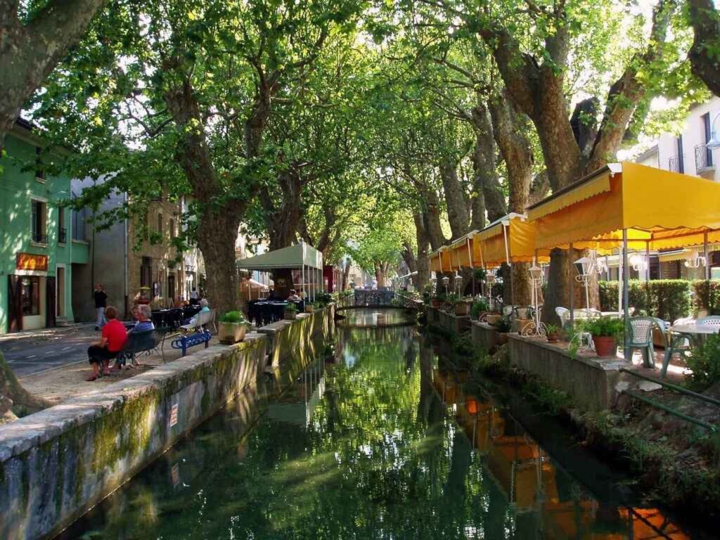 Canal bordé d'arbres avec terrasses de cafés aux stores jaunes et passants sur un banc dans un village pittoresque.