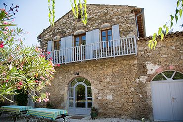 Mas provençal en pierre aux volets bleus avec balcon, terrasse et jardin fleuri sous le soleil.