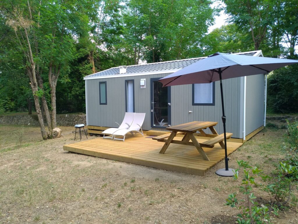 Mobil-home gris moderne avec terrasse en bois, table de pique-nique et transats sous un parasol en lisière de forêt.