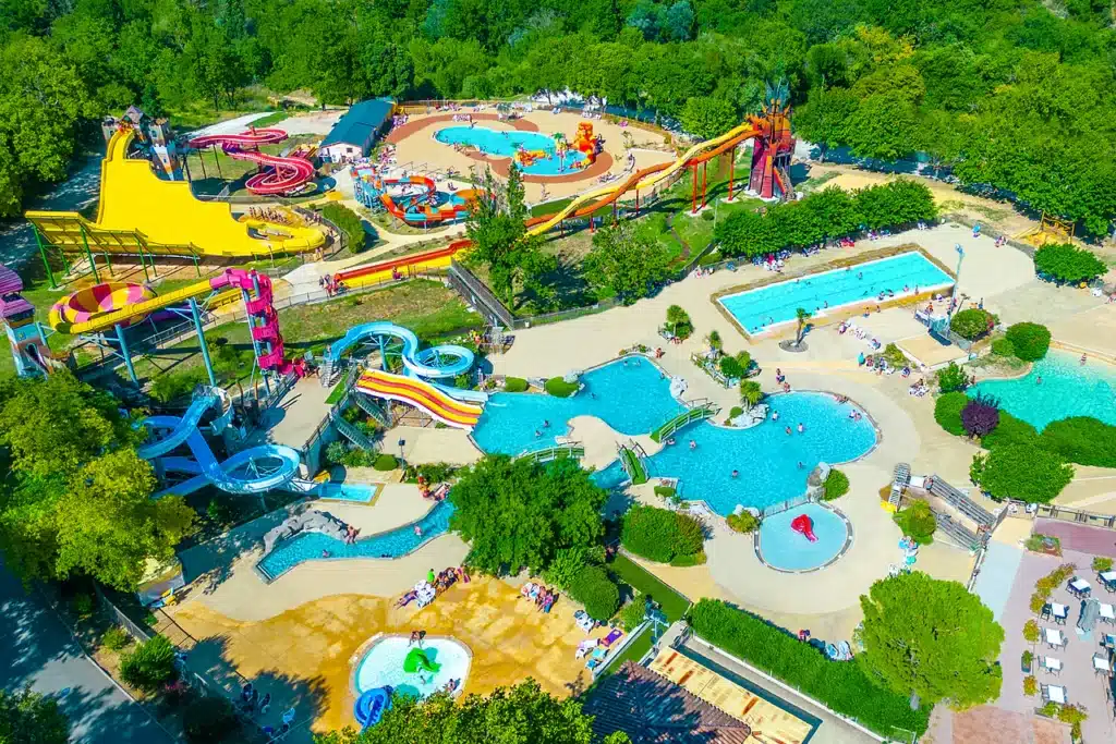 Vue aérienne d'un parc aquatique ensoleillé avec toboggans colorés, piscines lagon et espaces de détente arborés.