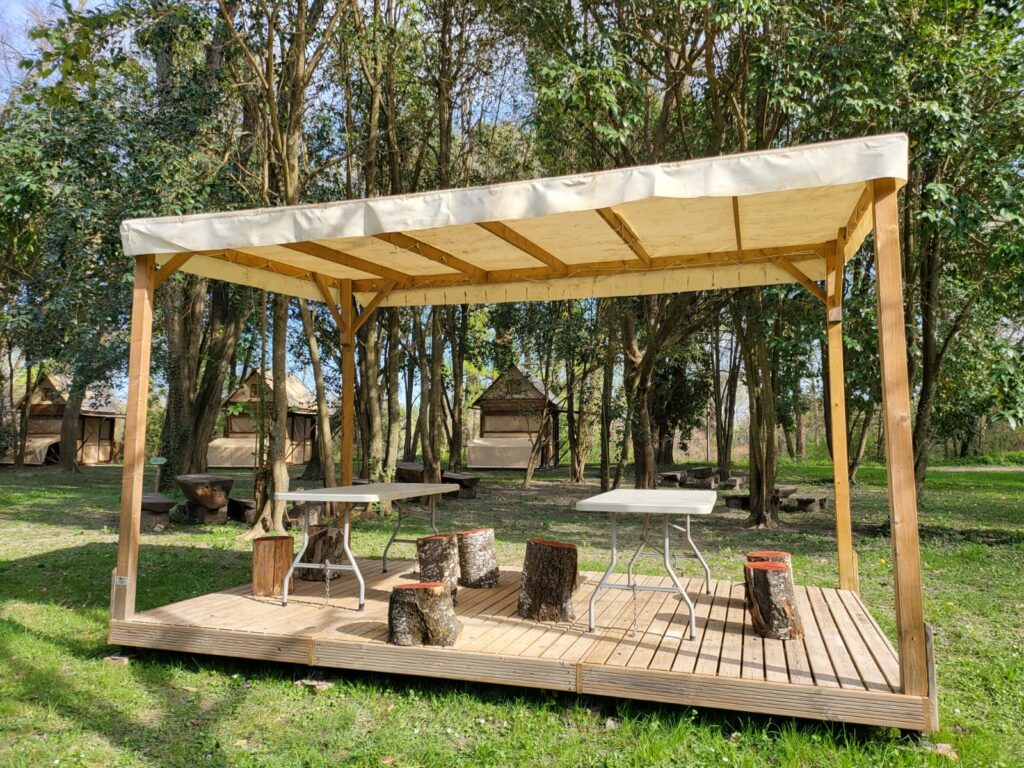 Pergola en bois avec terrasse, tables et tabourets en bûches dans un espace ombragé en forêt.