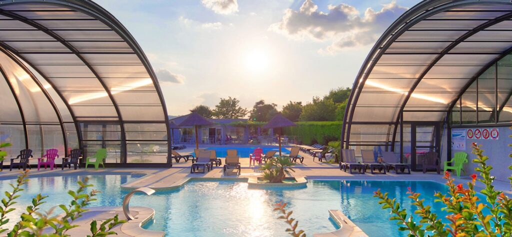 Complexe aquatique avec bassins couverts, transats et parasols sous un soleil couchant pour des vacances en camping.
