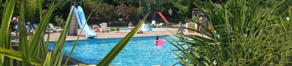 Piscine de camping avec toboggans bleu et rouge, vacanciers et chaises longues dans un cadre verdoyant.
