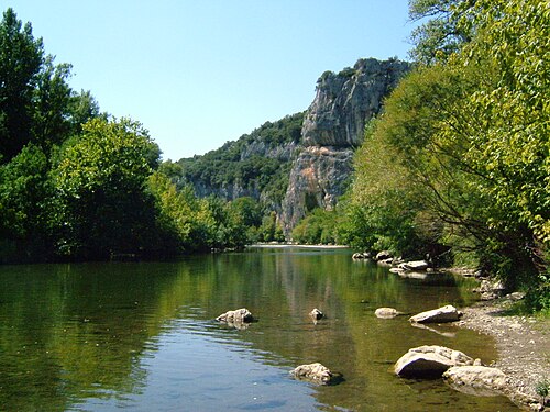 Rivière paisible entourée de forêts et de hautes falaises rocheuses sous un ciel bleu dégagé.