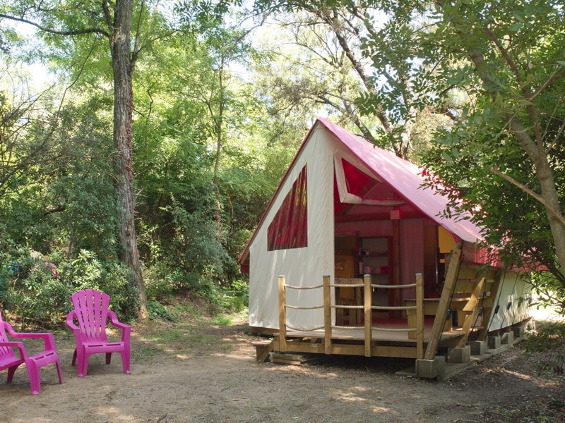 Tente glamping avec toit rose et terrasse boisée, deux chaises Adirondack roses dans un cadre naturel paisible.