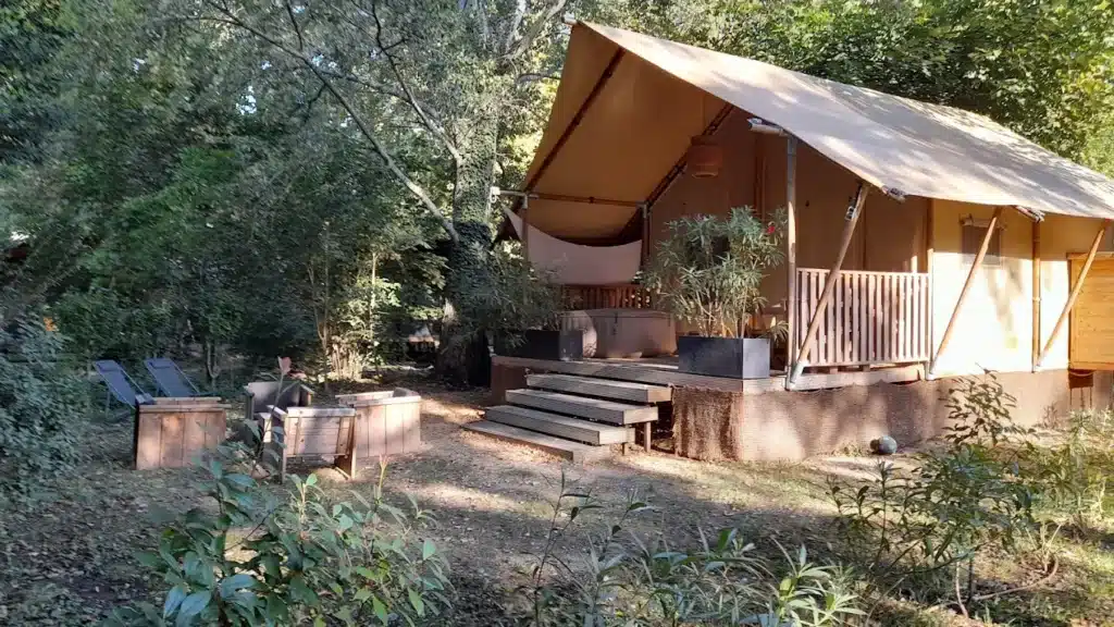 Tente safari de luxe avec terrasse en bois, jacuzzi et mobilier de jardin dans une forêt ensoleillée.