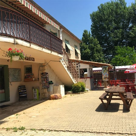 Réception d'un camping avec terrasse ombragée, tables de pique-nique en bois et mobilier rouge sous le soleil.
