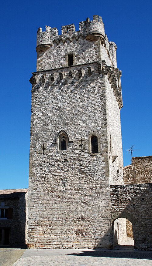 Haute tour médiévale en pierre claire avec créneaux et mâchicoulis sous un ciel bleu dégagé.