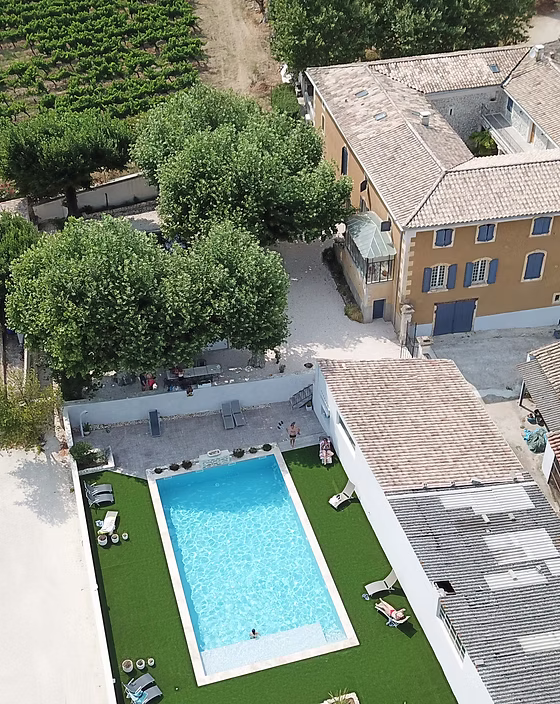 Vue aérienne d'une villa avec piscine rectangulaire, pelouse synthétique, grand bâtiment ocre et vignoble au loin.