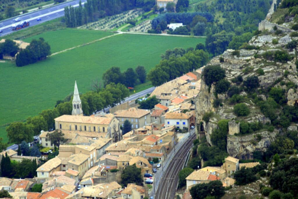 Village provençal niché au pied d'une falaise avec son église, des champs verts et une voie ferrée en contrebas.